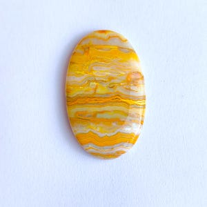 May include: Oval-shaped gemstone with a vibrant yellow and orange banded pattern. The polished stone has a smooth, glossy surface and a mix of colors, including white and gray, creating a layered effect. The gemstone is set against a white background.