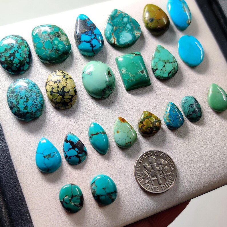Puede incluir: Una colecci&oacute;n de piedras preciosas de turquesa en varias formas y tama&ntilde;os, incluyendo ovaladas, en forma de l&aacute;grima y rectangulares. Las piedras muestran una gama de tonos azules y verdes con vetas oscuras. Se incluye una moneda de diez centavos estadounidense como referencia de tama&ntilde;o.