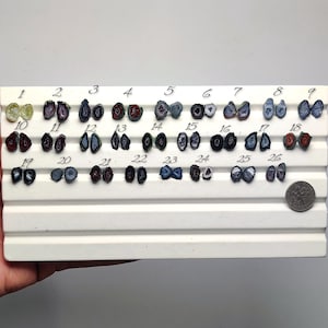 May include: A white display board featuring twenty-six pairs of small, polished agate slice earrings, each numbered from 1 to 26. The stones exhibit various colors including green, red, blue, black, and purple, with a US dime for scale.
