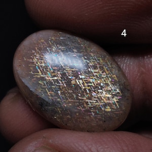 Puede incluir: Una gema pulida de forma ovalada con una superficie translúcida e iridiscente. La piedra presenta una mezcla de colores marrón, dorado e iridiscente, con un efecto brillante. El número "4" es visible en el fondo.
