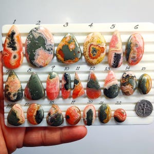 Puede incluir: Una colección de piedras preciosas pulidas en varias formas y colores, incluyendo rojo, naranja, rosa, verde y negro. Las piedras se muestran en una cuadrícula blanca, con una moneda de diez centavos estadounidense para comparar el tamaño.