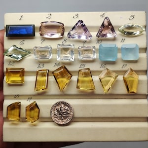 Puede incluir: Una colección de diecinueve piedras preciosas facetadas sueltas dispuestas en una bandeja de exhibición, numeradas del uno al diecinueve. Las piedras varían en forma, incluyendo cortes rectangulares, triangulares y marquesa, con colores que van desde el azul, rosa y amarillo hasta el transparente. Se incluye una moneda de diez centavos de EE. UU. para la escala.