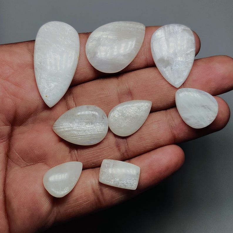 May include: A collection of eight polished, white gemstones in various shapes, including teardrop, oval, and rectangular. The stones have a smooth, translucent appearance, with subtle internal patterns. The gemstones are displayed on a hand, showcasing their natural beauty.
