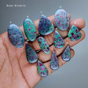 Könnte beinhalten: Eine Sammlung von Rubin-Kyanit-Edelsteinen in verschiedenen Formen, darunter oval und tropfenförmig. Die Steine zeigen eine Mischung aus grünen, blauen und roten Farbtönen. Der Text "RUBY KYANITE" ist oben im Bild zu sehen.