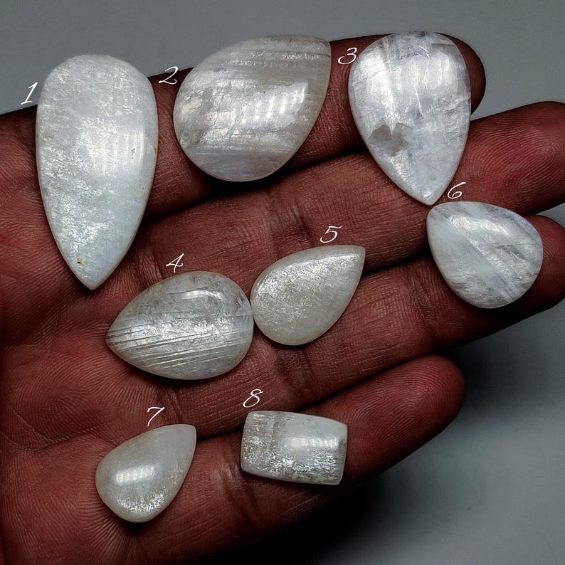 May include: A collection of eight polished, iridescent moonstone gemstones in various shapes, including teardrop, oval, and rectangular. The stones display a milky white color with subtle internal patterns and a pearlescent sheen. The gemstones are displayed on a dark surface.