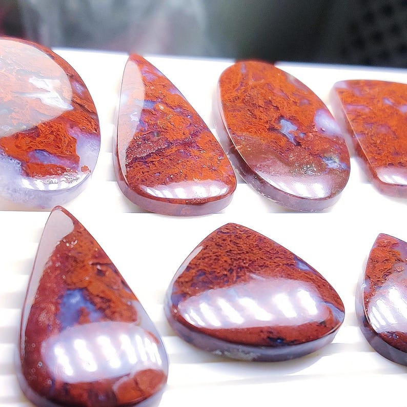 May include: A selection of polished gemstones in various shapes, such as oval and teardrop. The stones exhibit a deep red and brown color with blue undertones, and a glossy finish. The gemstones are presented against a white backdrop, showcasing their natural beauty.