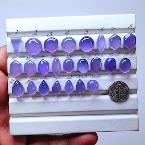 Puede incluir: Una colección de veinticuatro gemas pulidas de color lavanda, en diversas formas, incluyendo redondas, ovaladas y en forma de lágrima. Las gemas están dispuestas sobre una tabla blanca, con una moneda de diez centavos estadounidense para comparar el tamaño. Numeradas del 1 al 24.