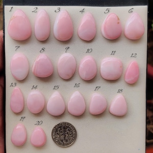 May include: A collection of twenty smooth, polished pink opal cabochons arranged on a display card, numbered one through twenty. A United States one dime coin is placed next to the twentieth stone for scale, showing the relative size of the gems.
