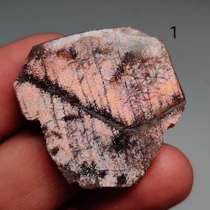 May include: A polished, iridescent gemstone with a mix of pink, brown, and black hues. The stone has a textured surface with a prominent dark vein running across it. The number "1" is visible in the upper right corner.