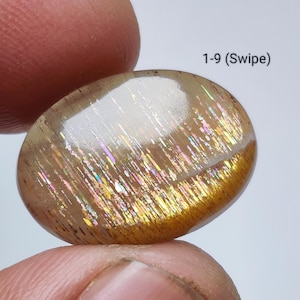 May include: Oval-shaped sunstone gemstone with a golden-brown hue and iridescent flashes. The stone is held between fingers, showcasing its polished surface and natural patterns. Text on the image reads "1-9 (Swipe)".