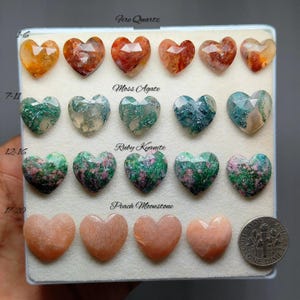 May include: A collection of heart-shaped gemstones arranged on a display board. Rows include Fire Quartz, Moss Agate, Ruby Kyanite, and Peach Moonstone. A US dime is placed next to the bottom row for scale, showing the relative size of the stones.
