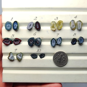 May include: Ten pairs of small, irregular geode slices arranged in rows on a white display board, numbered 1 through 10. Each pair features distinct colors including blue, red, yellow, grey, and black. A US dime is placed next to the tenth pair.