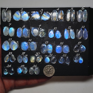 May include: A collection of twenty-six pairs of moonstone cabochons arranged on a black display board, each numbered from 1 to 26. The stones exhibit blue adularescence and varying shapes, including oval and teardrop. A US dime is included for scale.