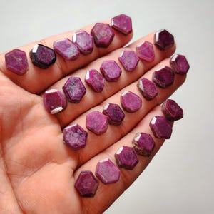 May include: A collection of numerous small, faceted, hexagonal-shaped ruby gemstones in varying shades of deep pink and red. Each individual stone features a flat top surface with angled sides, displaying natural inclusions and a semi-opaque texture.