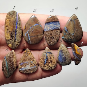 Pode incluir: Nove pedras preciosas de opala boulder polidas de várias formas, incluindo ovais e gotas, com matriz de ferro castanho e veios de cor azul e amarela iridescente, cada uma marcada com um pequeno número preto de um a nove.