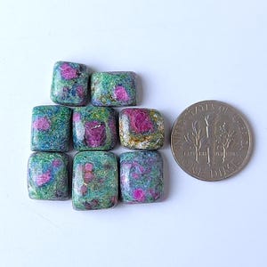 Op de afbeelding: Negen vierkant geslepen robijn-zoisiet edelstenen met een munt voor maatvergelijking. De edelstenen hebben een mix van roze, groene en witte kleuren. De edelstenen zijn gepolijst en hebben afgeronde randen.
