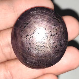 Puede incluir: Una gema grande, ovalada, de color rojo púrpura, con un patrón de estrella de seis puntas. La superficie tiene un aspecto texturizado con inclusiones oscuras y estrías visibles. La gema se sostiene en una mano, mostrando su tamaño y color.