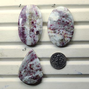 May include: Three polished gemstones in white, pink, and grey hues. Two are oval, one is teardrop-shaped. A coin is included for scale. The gemstones display a mix of colours and textures.