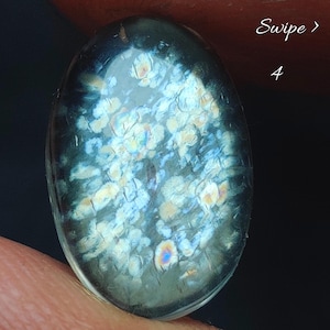 May include: An oval-shaped cabochon gemstone featuring a dark, translucent base with internal inclusions that create a shimmering, iridescent pattern of white, blue, and gold flecks. The surface is smooth and polished, reflecting light across its dome.