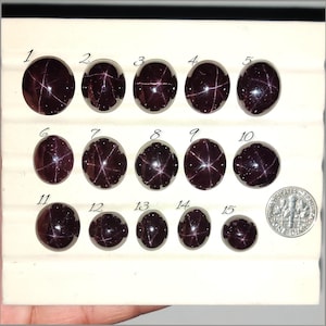 May include: Fifteen dark red gemstones of varying sizes, each with a star-like pattern, are displayed. A US dime is included for size comparison. The gemstones are numbered 1 through 15, showcasing their unique characteristics.