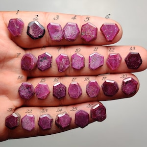 Può includere: Una collezione di ventisei rubini sfaccettati, dal rosa intenso al rosso, disposti in file su un palmo. Ogni gemma è numerata da 1 a 26 con inchiostro nero. Le pietre presentano varie forme geometriche esagonali e irregolari con inclusioni naturali visibili e superfici levigate.