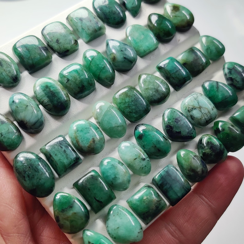 May include: A collection of polished, cabochon-cut emerald gemstones arranged in rows on a white display tray. The stones exhibit various shades of green, ranging from light mint to deep forest tones, with natural inclusions and smooth, glossy surfaces.