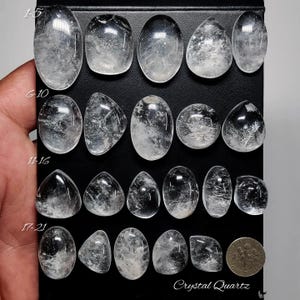 May include: A collection of twenty-one clear, polished crystal quartz cabochons in various shapes including oval, round, and teardrop. The stones are arranged in rows on a black display board, with a US dime coin for scale next to the last row.