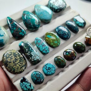 Op de afbeelding: Een verzameling gepolijste turkoois cabochons in diverse vormen en maten, met opvallende matrixpatronen in blauwe, groene en bruine tinten, gerangschikt in rijen op een lichtgrijs presentatieplateau met individuele vakjes voor elke steen.