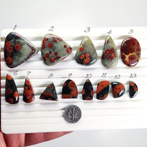 May include: A collection of polished gemstones in various shapes, including teardrop, oval, and triangular. The stones feature a mix of colors, including red, black, and green, with a dime for size comparison. The gemstones are arranged on a white surface.