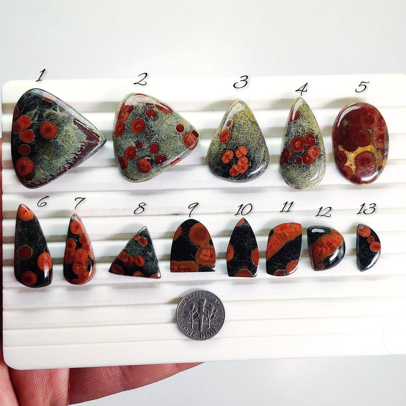 May include: A collection of polished gemstones in various shapes, including teardrop, oval, and triangular. The stones feature a mix of colors, including red, black, and green, with a dime for size comparison. The gemstones are arranged on a white surface.