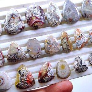 May include: A collection of polished agate gemstones in various shapes and sizes. The stones display a range of colors, including white, brown, red, and gray, with intricate patterns and banding. The gemstones are displayed on a white surface.