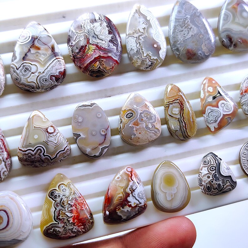 May include: A collection of polished agate gemstones in various shapes and sizes. The stones display a range of colors, including white, brown, red, and gray, with intricate patterns and banding. The gemstones are displayed on a white surface.