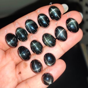 May include: Thirteen oval-shaped black star diopside gemstones, each featuring a distinct four-rayed star effect. The stones are numbered one through thirteen in white script, showcasing a polished, reflective surface with deep dark green tones.