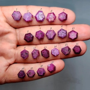 May include: A collection of twenty small, faceted, hexagonal-shaped pinkish-red rubies arranged in rows. Each gemstone is individually numbered from 1 to 20 with a small black digit placed directly above it, showcasing natural variations in colour.