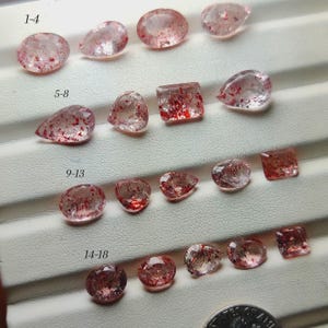 Puede incluir: Colección de gemas rosas translúcidas con inclusiones rojas, talladas en diversas formas, como óvalos, peras y cuadrados. Las piedras están dispuestas sobre una superficie blanca. Se incluye una moneda como referencia de tamaño.