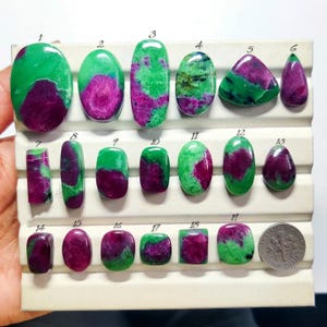 May include: A collection of nineteen polished ruby in zoisite cabochons arranged on a white display tray, numbered one through nineteen, with a United States dime coin included for scale to show the relative size of the gemstones.