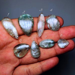 Puede incluir: Una colección de ocho piedras preciosas pulidas de varias formas, incluyendo ovaladas y en forma de lágrima. Las piedras muestran una mezcla de tonos claros, verdes y plateados, con patrones internos. Las piedras preciosas se sostienen en una mano.
