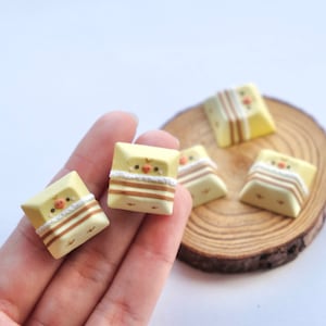 May include: Four yellow and brown keycaps with a cute cartoon chick design. The keycaps are on a wooden surface and a hand is holding two of them.