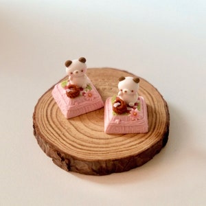 May include: Two small white figurines of animals sitting on pink cushions with brown accents. The cushions are decorated with small pink flowers. The figurines are sitting on a wooden base.