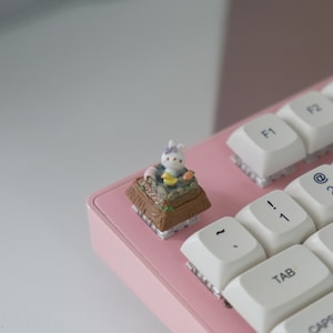 Può includere: Una tastiera rosa con tasti bianchi e una piccola figurina di coniglio sul tasto 'Esc'. La figurina è seduta in una piccola scena da giardino con una base marrone.