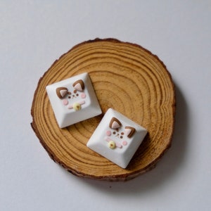 May include: Two white keycaps with brown ears and pink cheeks, resembling a cartoon cat, are displayed on a wooden surface.