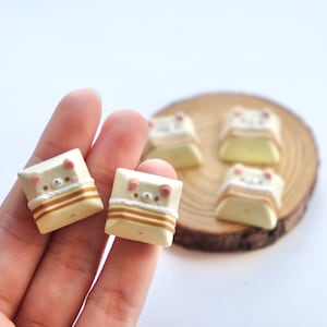 May include: Five small, yellow keycaps with a brown and white striped design and a cartoon bear face with pink cheeks. The keycaps are on a wooden surface and in a hand.