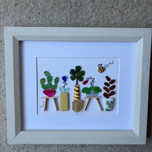 May include: A framed wall art piece featuring a collection of five potted plants and a bee created from colorful sea glass and small wooden sticks. The plants are arranged on a white background within a light-colored wooden frame. The artist signature Pam is visible.