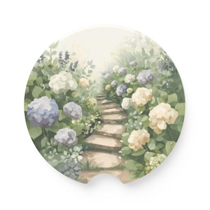 May include: A round car coaster featuring a watercolor painting of a stone path winding through a garden. The garden is filled with blue, white, and yellow hydrangea flowers and lush green foliage.