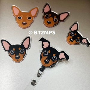 Op de afbeelding: Vier cartoon-hondenkopbadges, twee bruin en twee zwart, met roze oren. Een zwart hondenkopbadge is bevestigd aan een transparante, intrekbare badgehouder van plastic met een zwarte clip en zilveren metalen ringen.