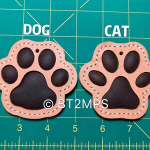 May include: Two leather paw print keychains, one labeled "DOG" and the other "CAT". The paw prints are brown with a tan outline and white stitching.
