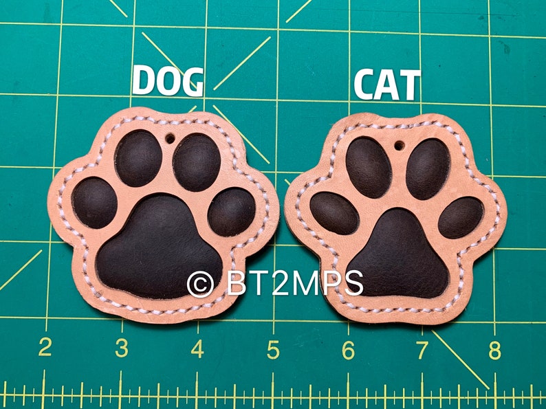 May include: Two leather paw print keychains, one labeled "DOG" and the other "CAT". The paw prints are brown with a tan outline and white stitching.