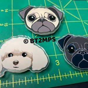 May include: Three acrylic dog head keychains. One is a white poodle, one is a light brown pug, and one is a black pug.