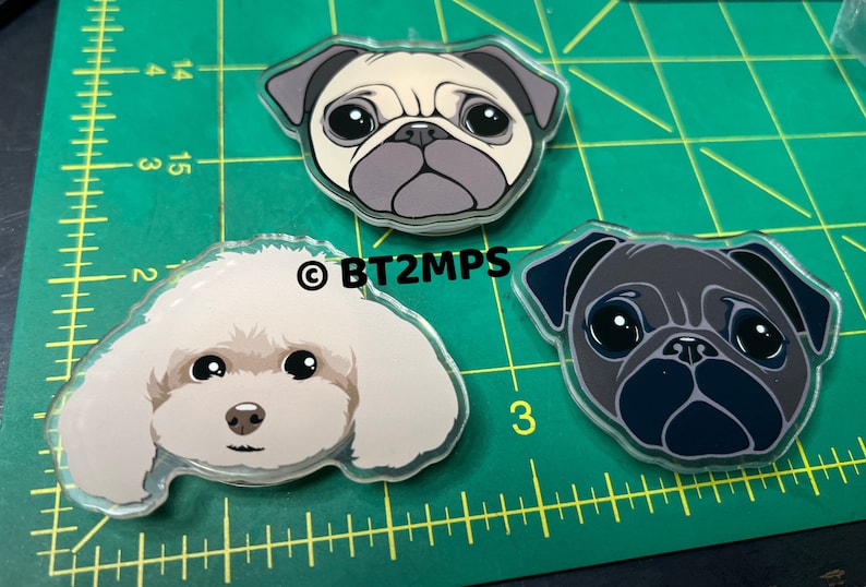 May include: Three acrylic dog head keychains. One is a white poodle, one is a light brown pug, and one is a black pug.
