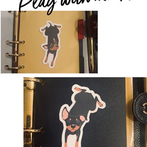 Puede incluir: Una pegatina de perro negro y marr&oacute;n con el texto "Play with me!!!" sobre un fondo amarillo. La pegatina est&aacute; en una agenda negra.